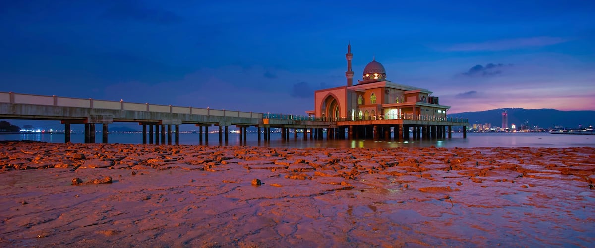 A mosque with a dome and minaret, a bridge, and a rocky shoreline at dusk.