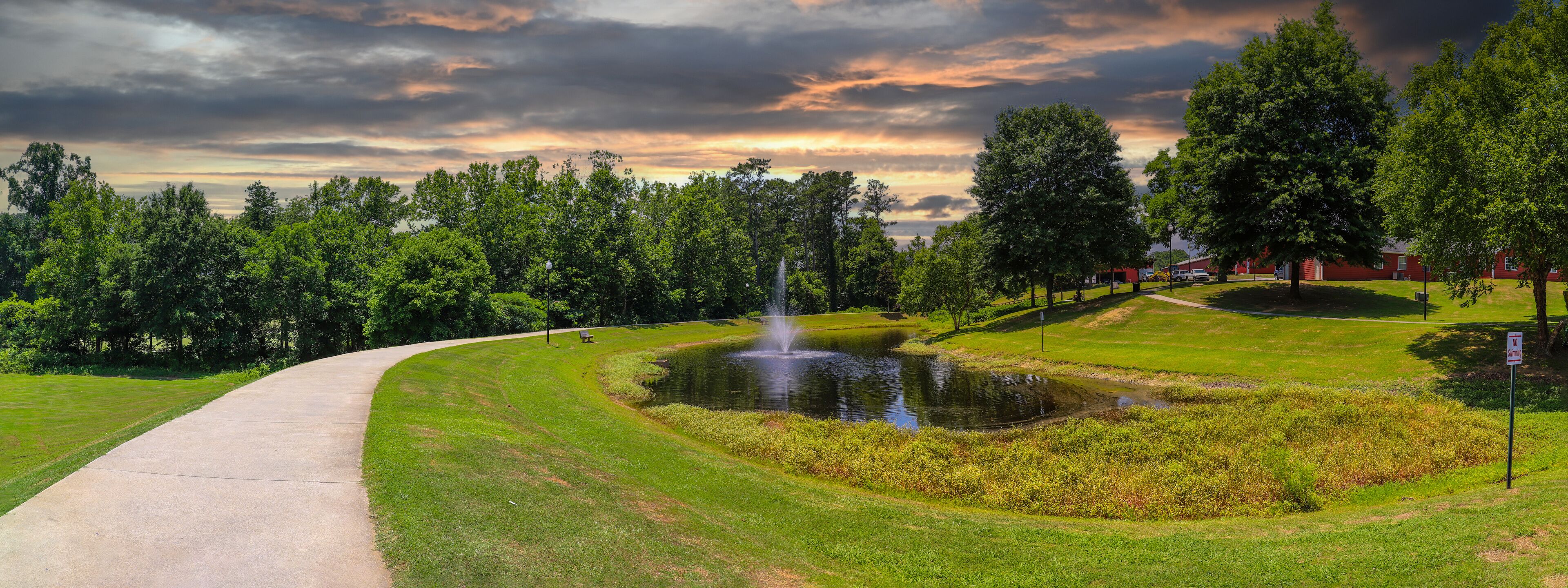 a panoramic shot of a gorgeous summer landscape in the park with a pond with a water fountain in the center surrounded by lush green trees, grass and plants with powerful clouds at sunset