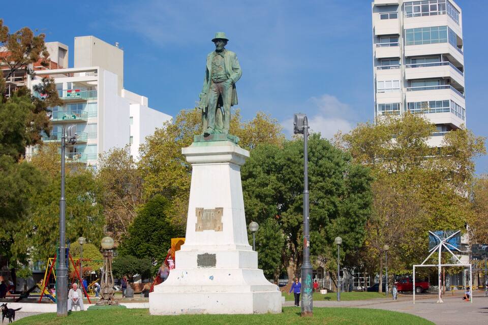 A statue of a man in a suit and hat on a pedestal in a park with trees and buildings in the background.