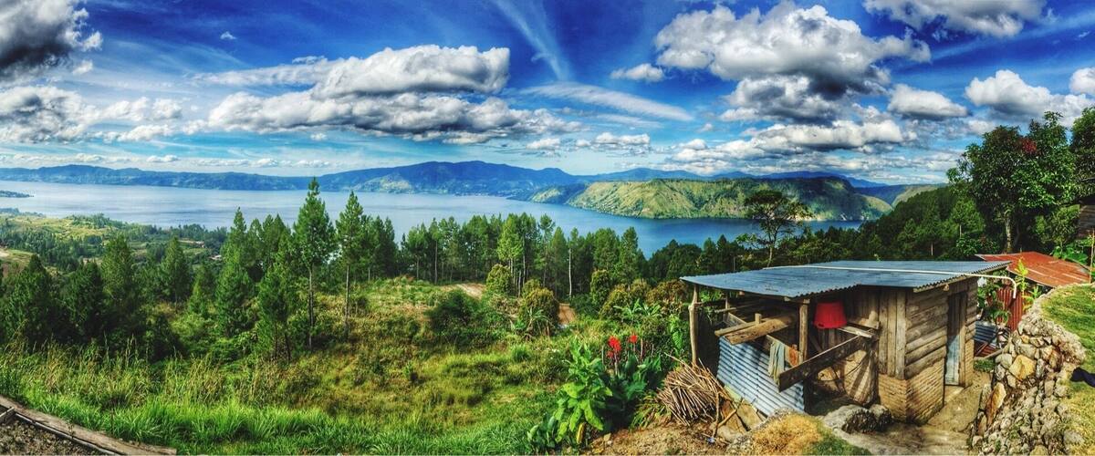 A rural landscape with a wooden cabin, a small shed, and a view of a lake surrounded by hills and trees.
