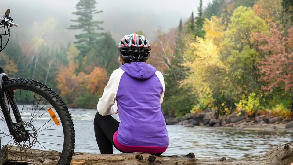 sitting female bicyclist admires beautiful autumn landscape in foggy morning during halt at sand beach of riviere-rouge river by corridor aerobique bicycle trail in province of quebec, canada