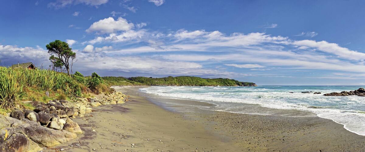 A sandy beach with waves, a rocky shoreline, and a forested hill in the background.