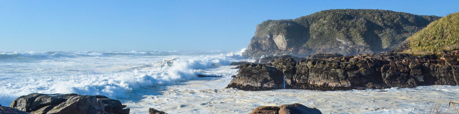 Una costa rocosa con olas rompiendo contra la orilla y un acantilado destacado al fondo.