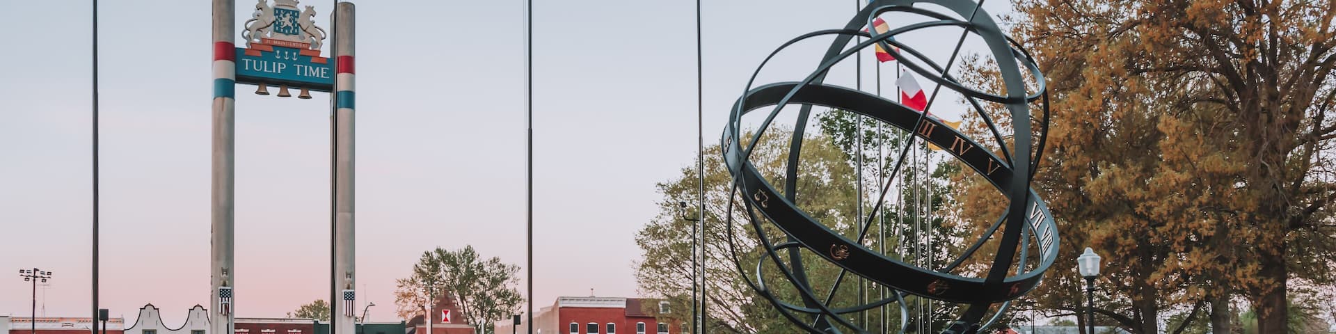 Multicolored spring tulips line a brick walkway towards the sundial and Tulip Toren Monument in Pella's Central Park at sunrise surrounded by crabapple trees, beds of tulips and flags flapping.