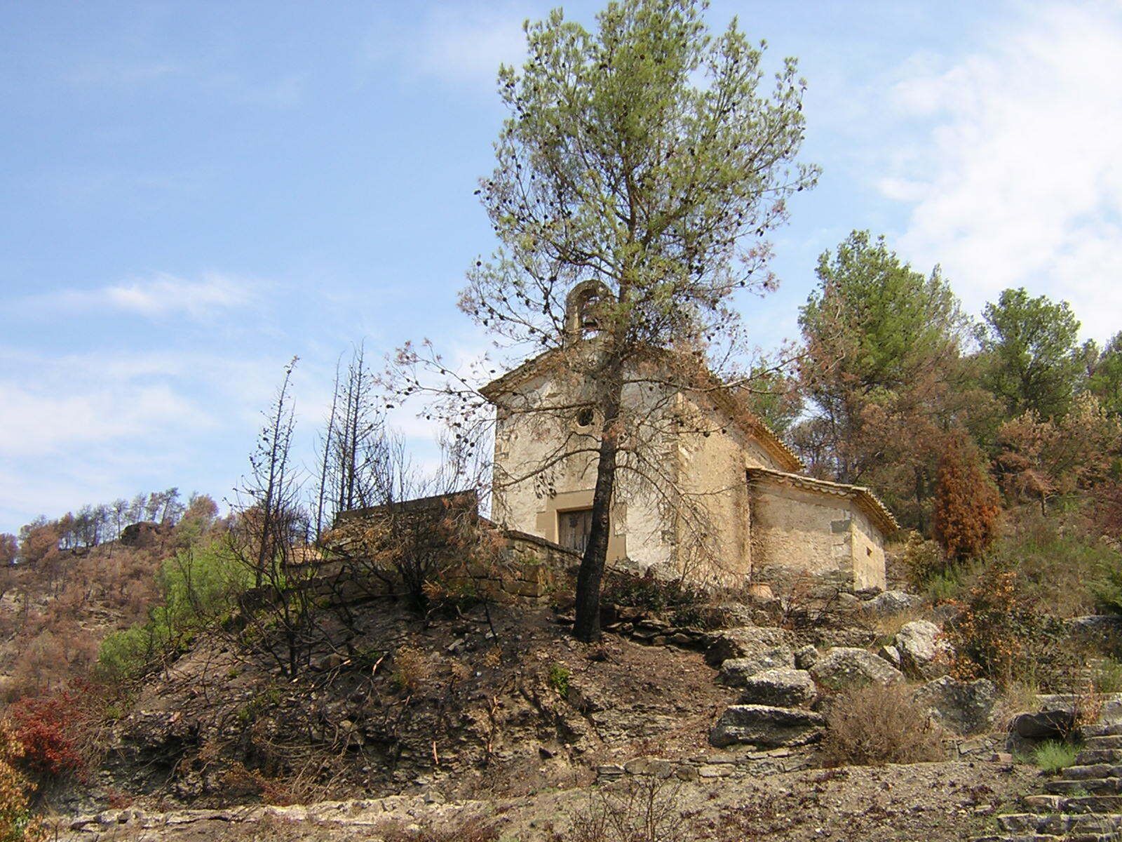 A stone chapel with a tree in front, surrounded by rocky terrain and sparse vegetation.
