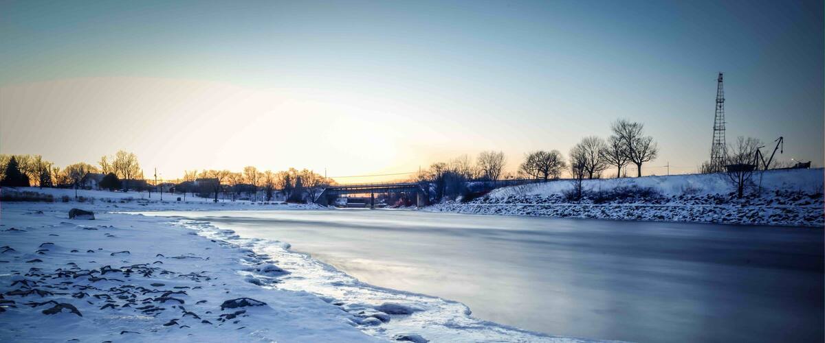 A frozen river with snow-covered banks and a bridge in the background.