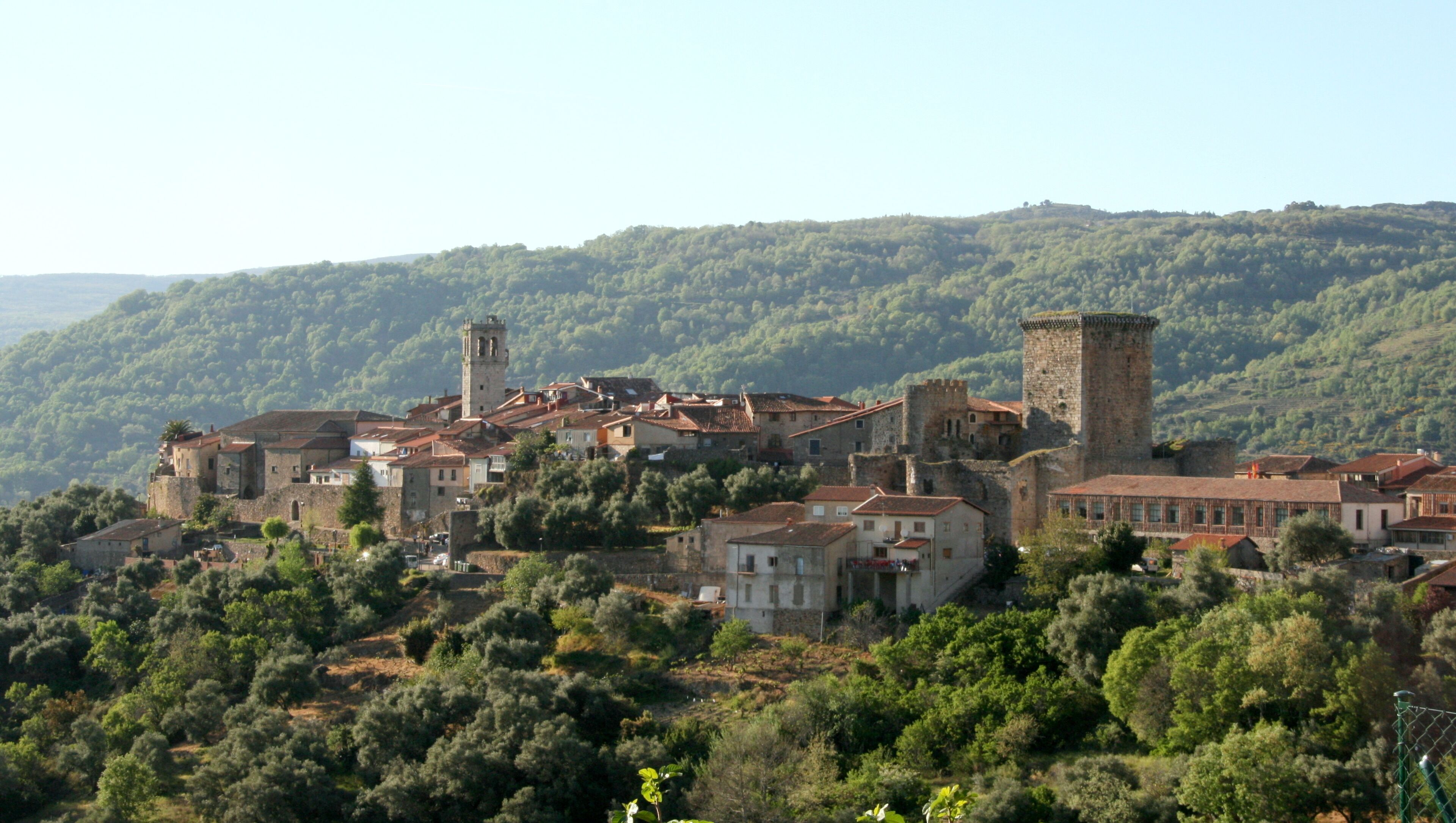 A historic town with medieval architecture, including towers and fortified walls, nestled in a hilly, forested landscape.