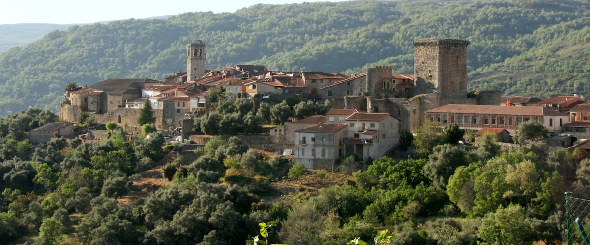 A historic town with medieval architecture, including towers and fortified walls, nestled in a hilly, forested landscape.