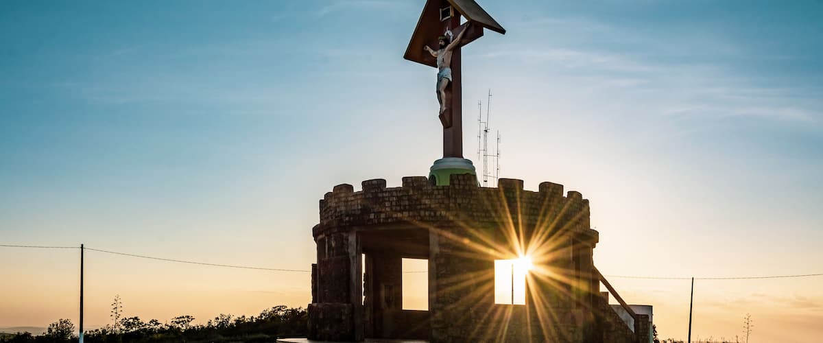 A statue of Jesus Christ on a cross, a sun setting in the background, and a paved walkway leading to the statue.