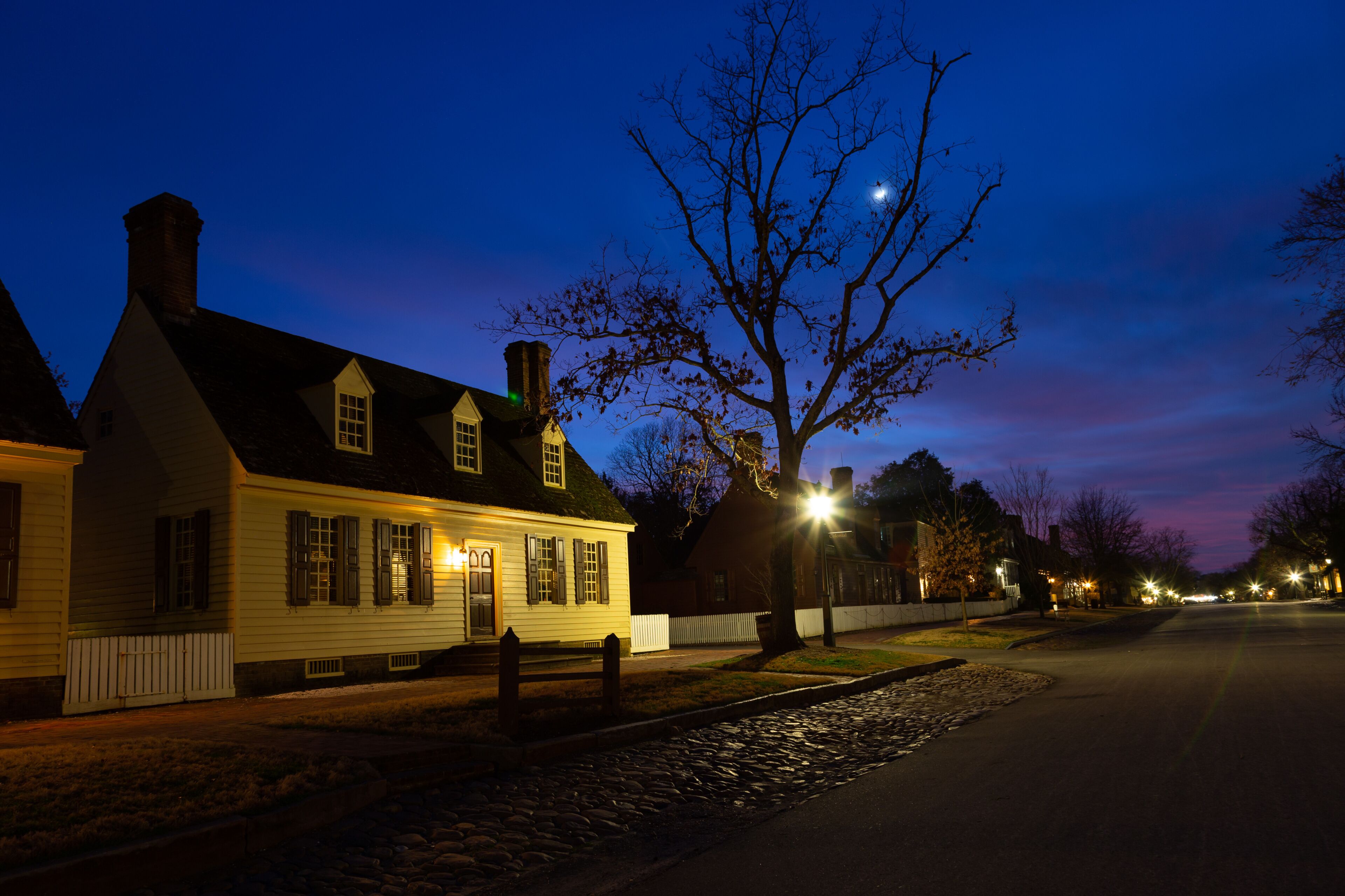 A historic white house with a dark roof, a wooden fence, and a tree with bare branches.