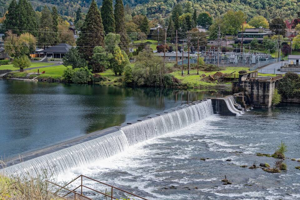 A dam with a waterfall, surrounded by greenery and buildings.