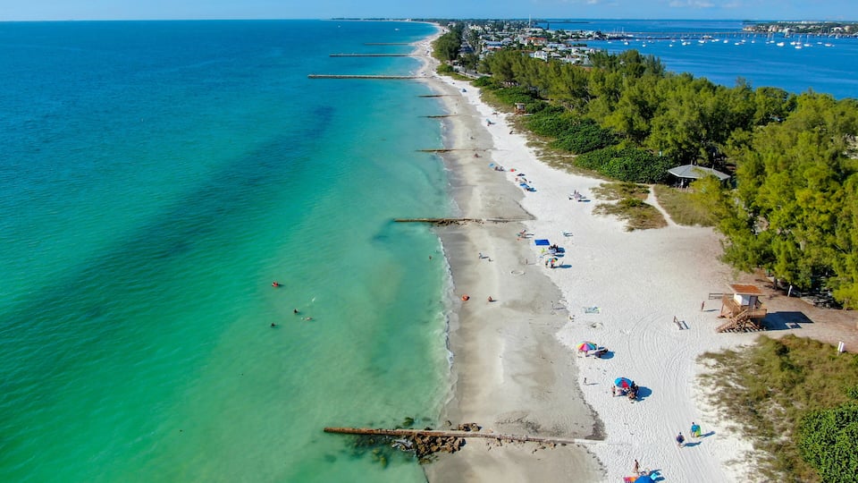 A coastal beach with clear turquoise waters, a sandy shore, and a line of trees along the beachfront.
