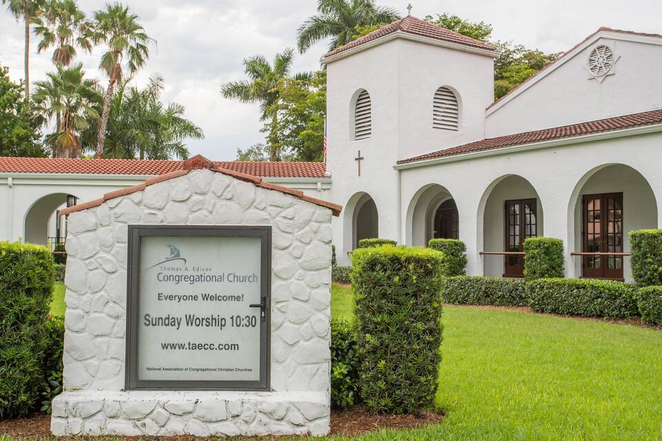 A white church with a stone signboard, surrounded by well-maintained hedges and palm trees.