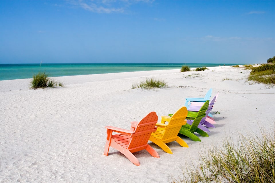Een strand met wit zand en turquoise water. Er staan kleurrijke Adirondack-stoelen op een rij in het zand.