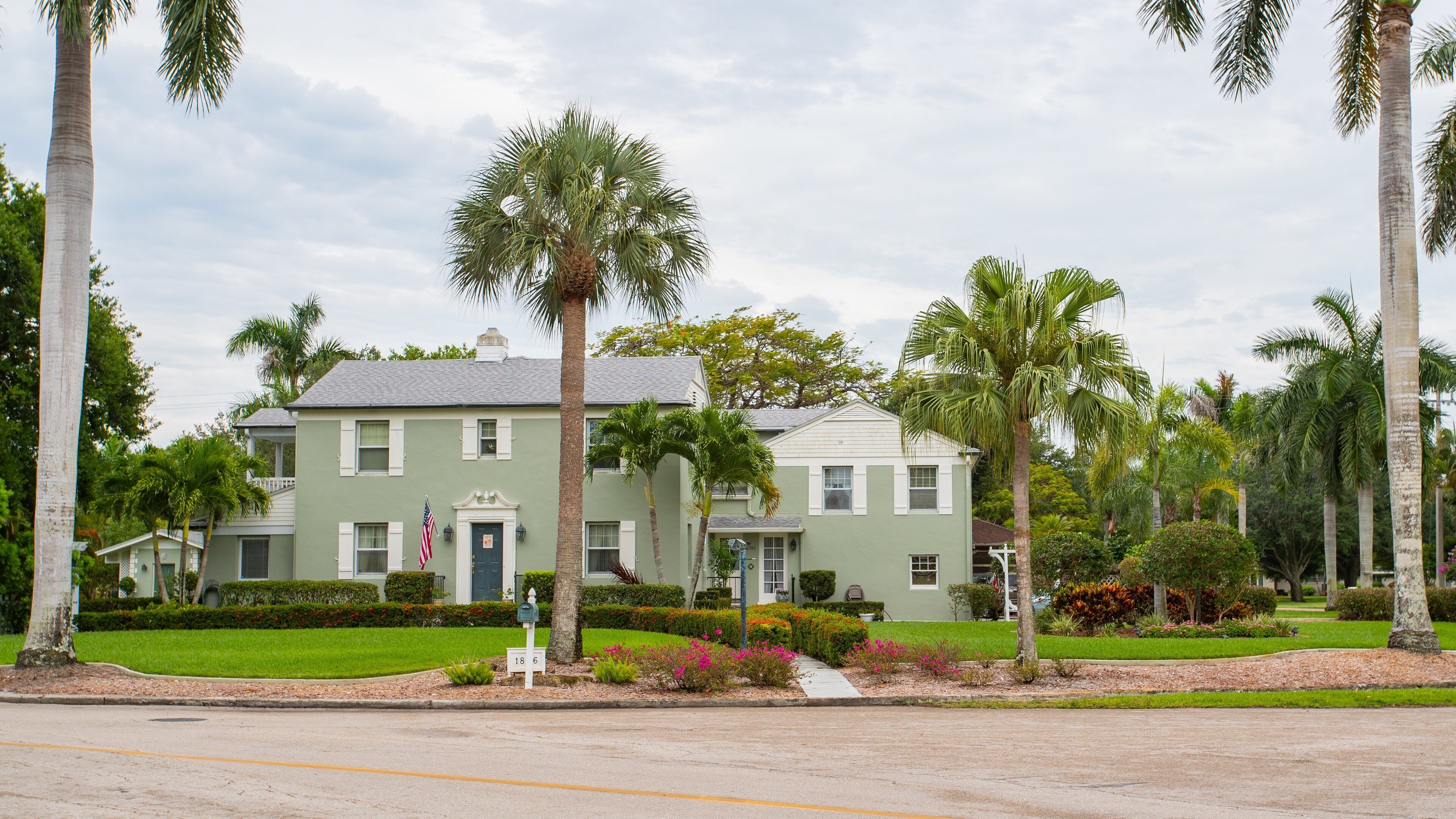 A green two-story house with a white porch, surrounded by palm trees and well-maintained gardens.