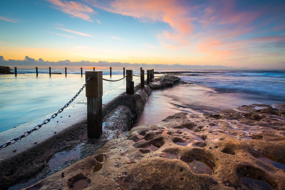 Een strand met een houten omheining en kettingafsluiting, rotsachtig terrein en een zonsondergang.