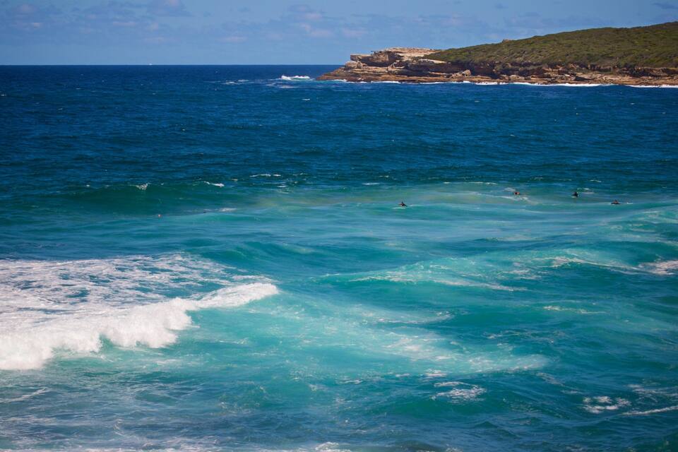 A coastal scene with clear turquoise waters, waves, and a rocky cliff with sparse vegetation.