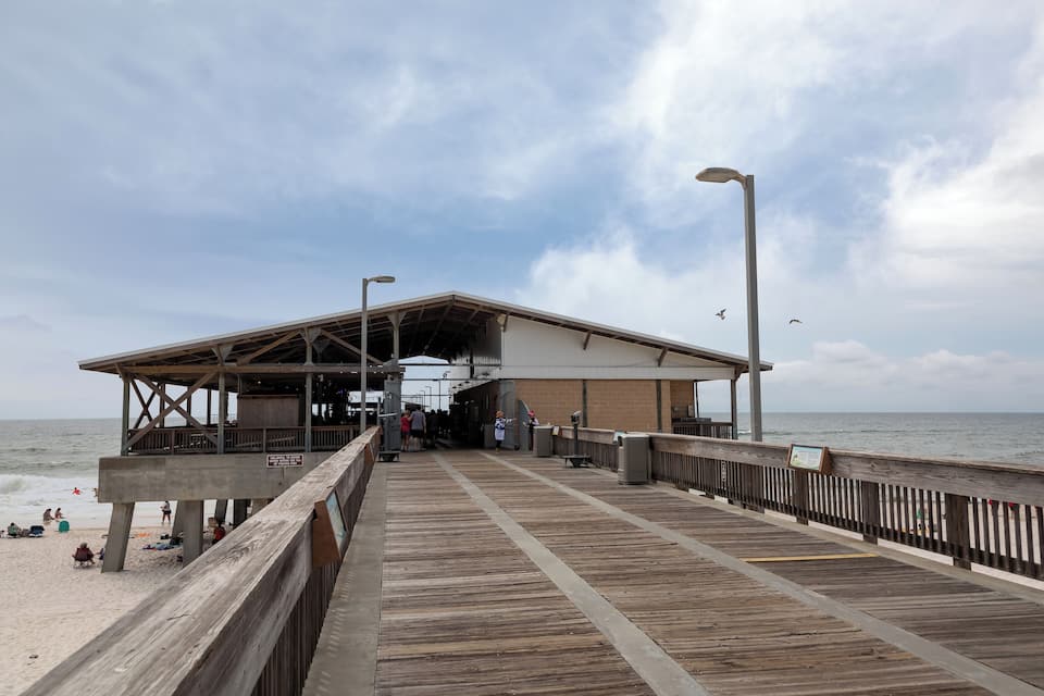 Un muelle de madera que conduce a una estructura en la playa con techo, una playa arenosa y el océano al fondo.