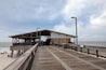 Een houten steiger die naar een gebouw aan het strand leidt, met een zandstrand en de oceaan op de achtergrond.