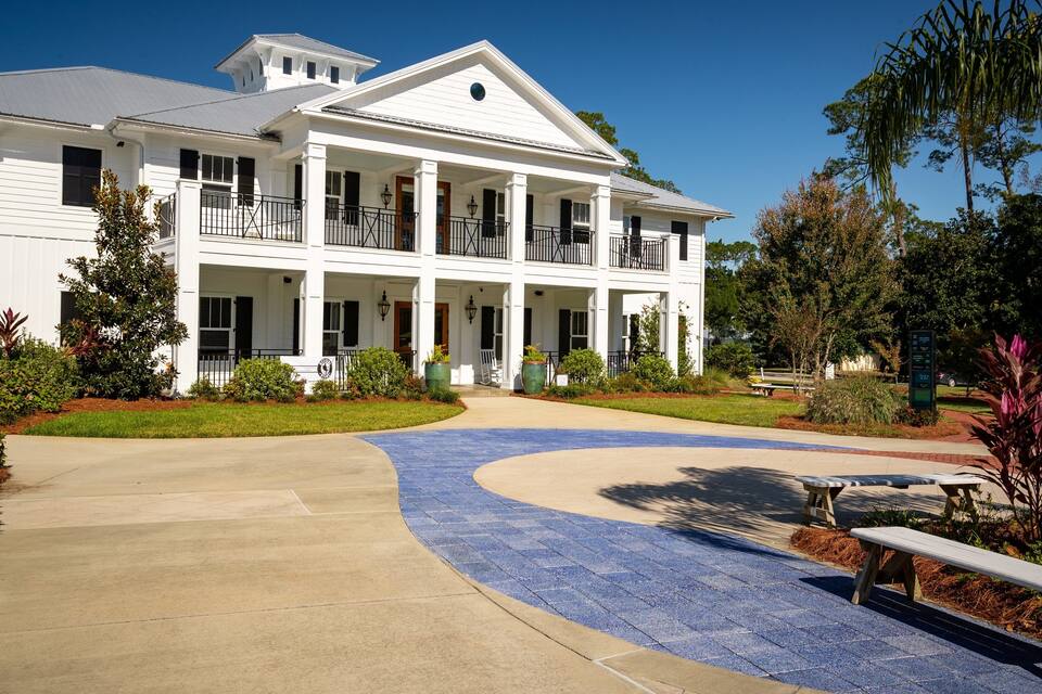 A white colonial-style house with a wrap-around porch, surrounded by well-maintained landscaping and a curved pathway.