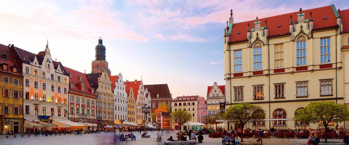Una plaza histórica europea con pavimento de adoquines, edificios tradicionales y una torre destacada.