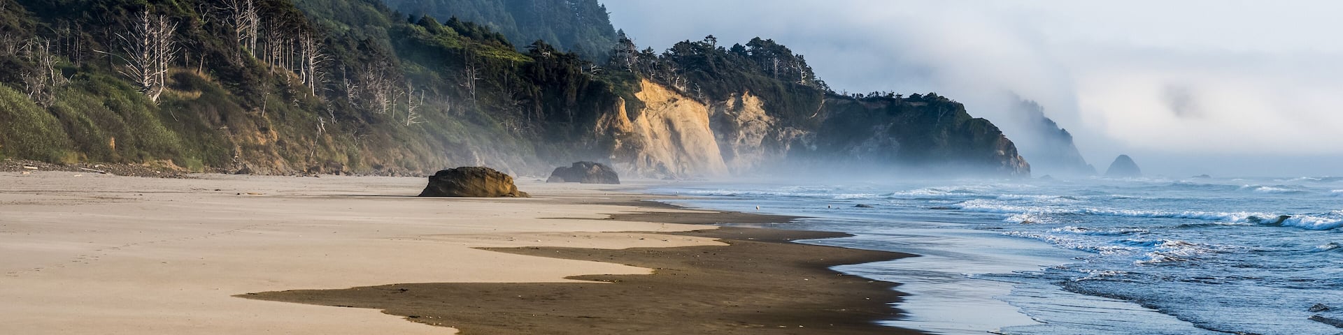 Una playa con un promontorio rocoso al fondo, rodeada de colinas boscosas.