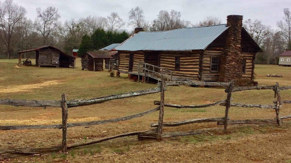 A rustic log cabin with a stone chimney, surrounded by a wooden fence and other wooden structures in a rural setting.