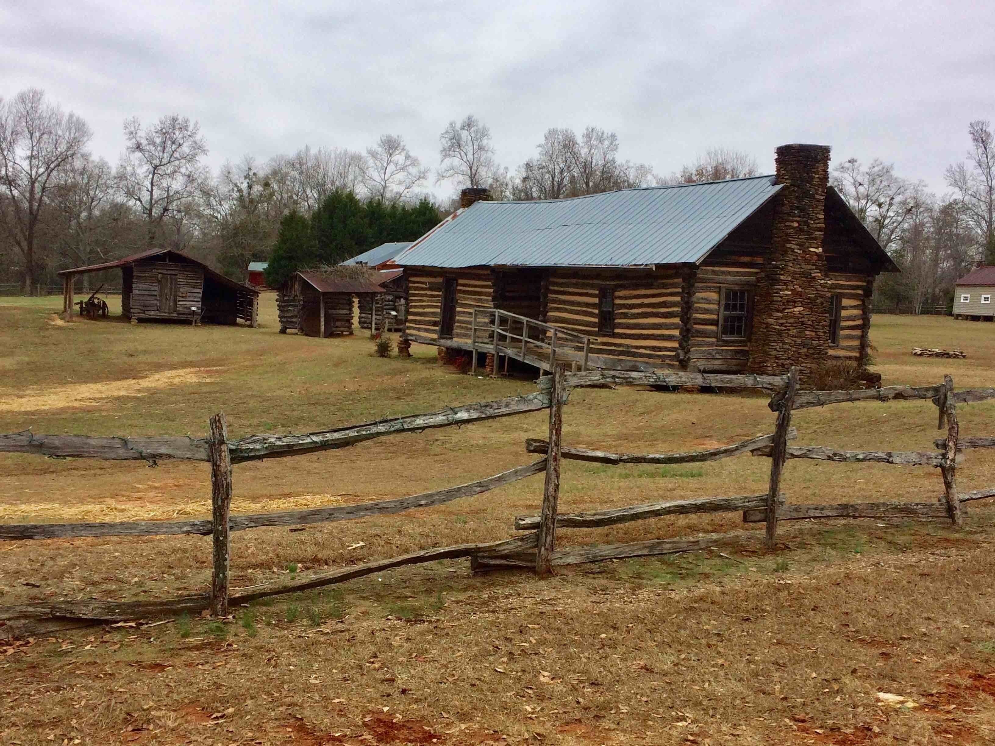 A rustic log cabin with a stone chimney, surrounded by a wooden fence and other wooden structures in a rural setting.