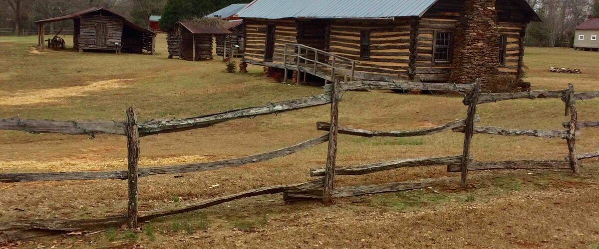 A rustic log cabin with a stone chimney, surrounded by a wooden fence and other wooden structures in a rural setting.