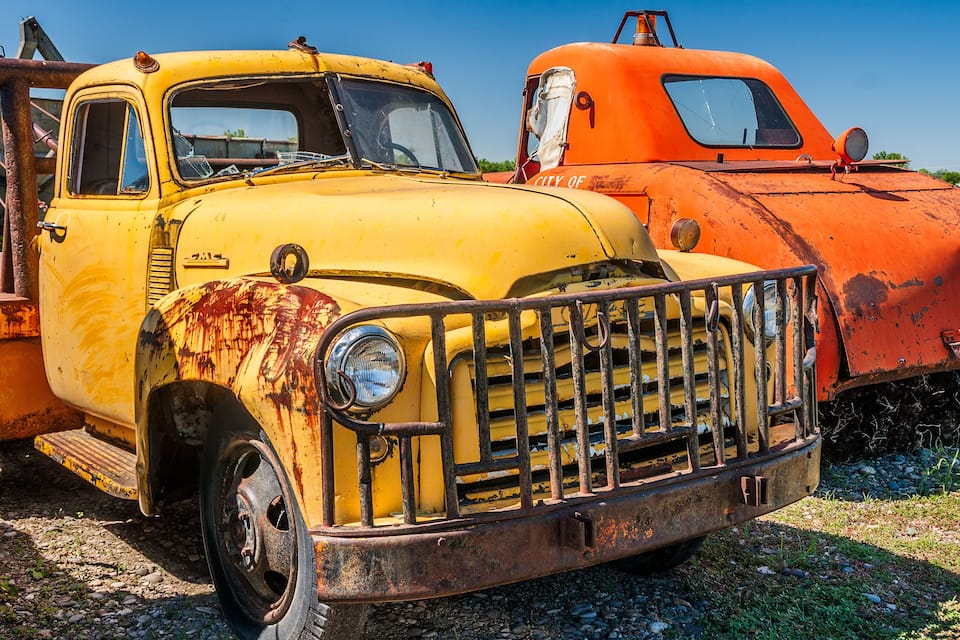 Un camion jaune rouillé, dont la calandre avant est endommagée, est garé à côté d’un autre camion, également rouillé, mais de couleur orange.