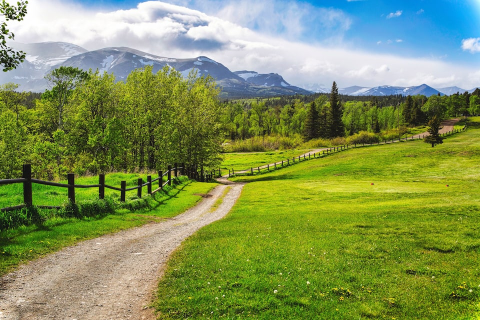 Un sentier de terre traverse un champ verdoyant, bordé d’une clôture en bois, et est entouré d’arbres, avec des montagnes en arrière-plan.