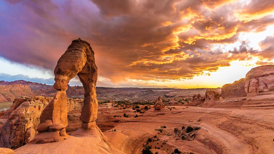 A rock arch formation in a desert landscape with a sunset sky.