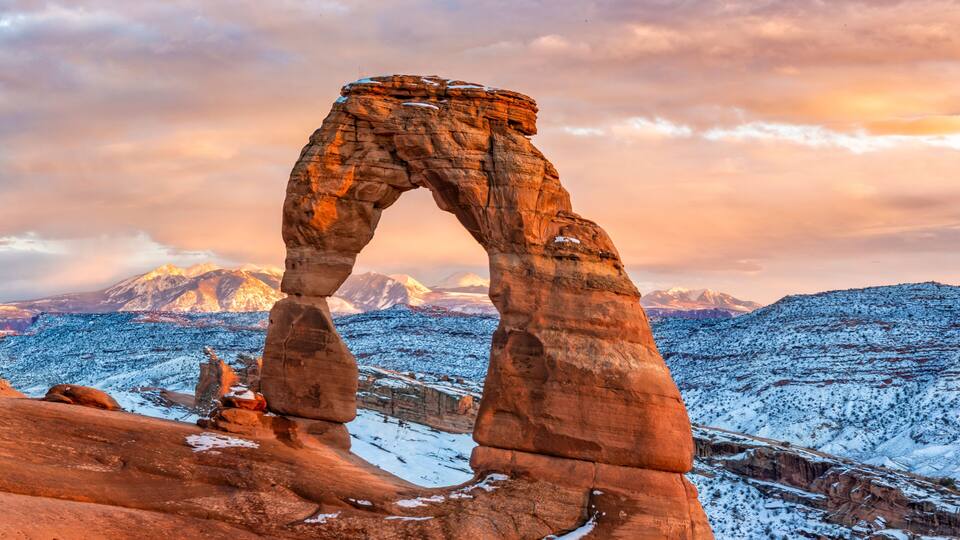 A rock arch formation with snow-covered terrain and mountains in the background.