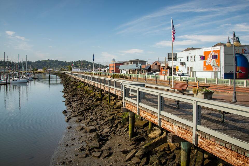 A waterfront promenade with a wooden walkway, boats docked on the left, and a building with a Coca-Cola sign on the right.