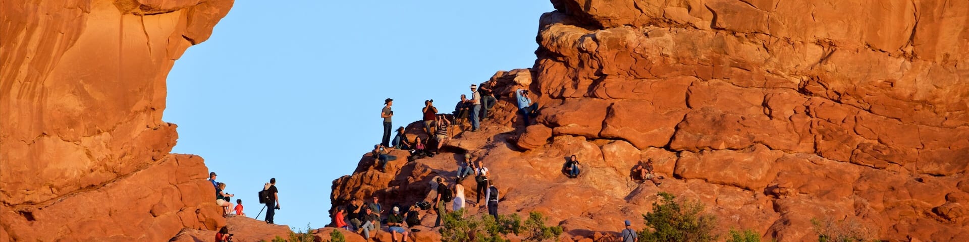 A large red rock arch with people climbing and sitting around it.