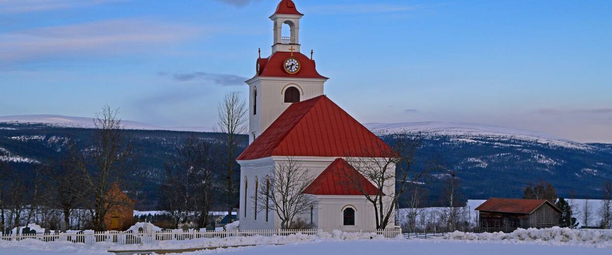 A church with a red roof and white walls, surrounded by snow-covered fields and mountains.