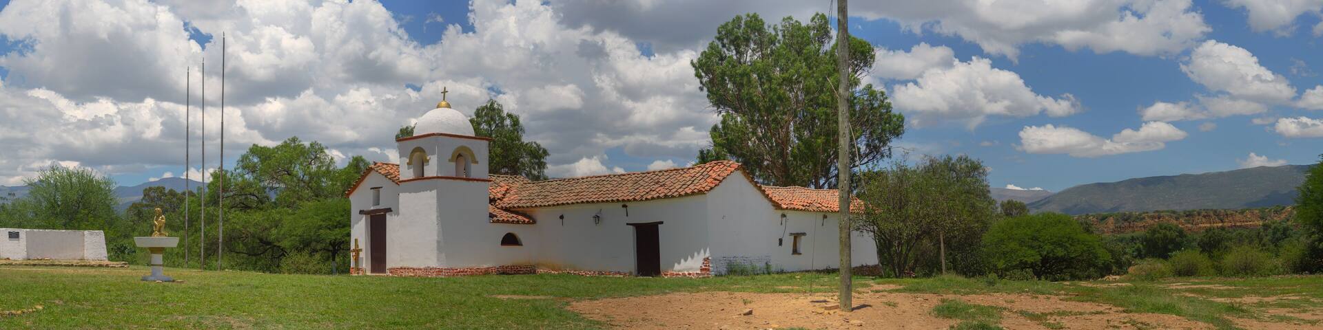 A white church with a red-tiled roof and a bell tower, surrounded by green grass and trees, with mountains in the background.