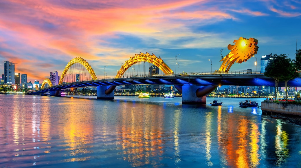 A bridge with two large, illuminated arches over a river, with city buildings in the background.