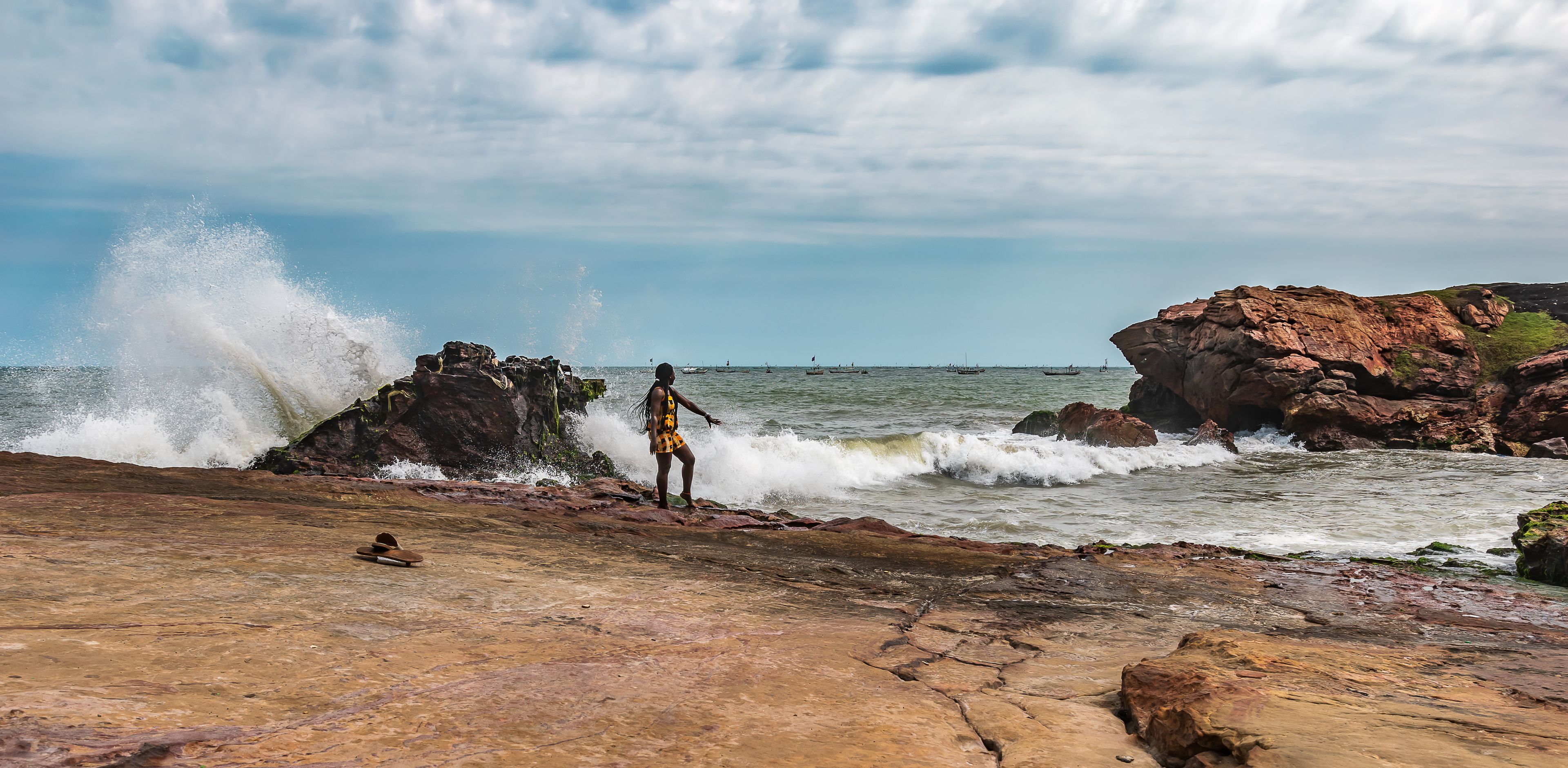 Uma costa rochosa com ondas quebrando nas pedras, uma pessoa em pé sobre elas, e barcos na água.