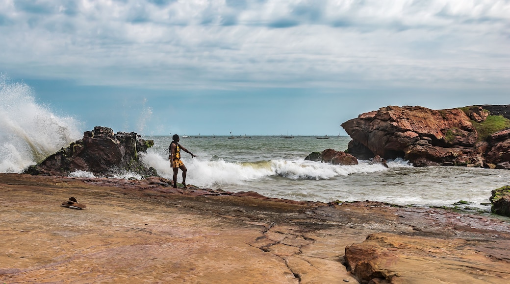 Uma costa rochosa com ondas quebrando nas pedras, uma pessoa em pé sobre elas, e barcos na água.