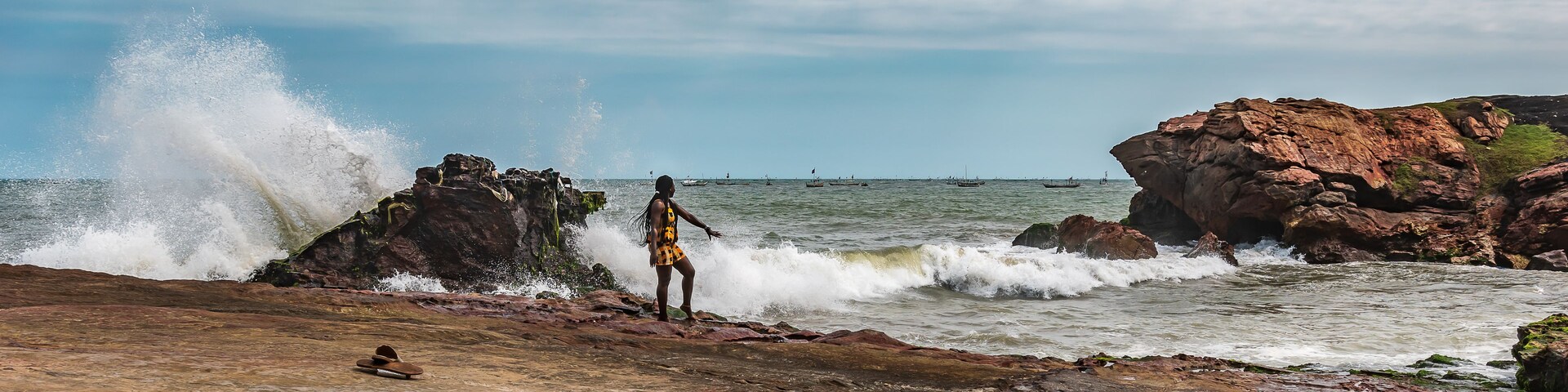 A rocky shoreline with waves crashing against the rocks, a person standing on the rocks, and boats in the water.