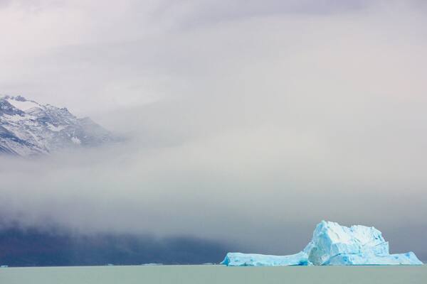A glacier with a large iceberg in a body of water, surrounded by mountains and clouds.