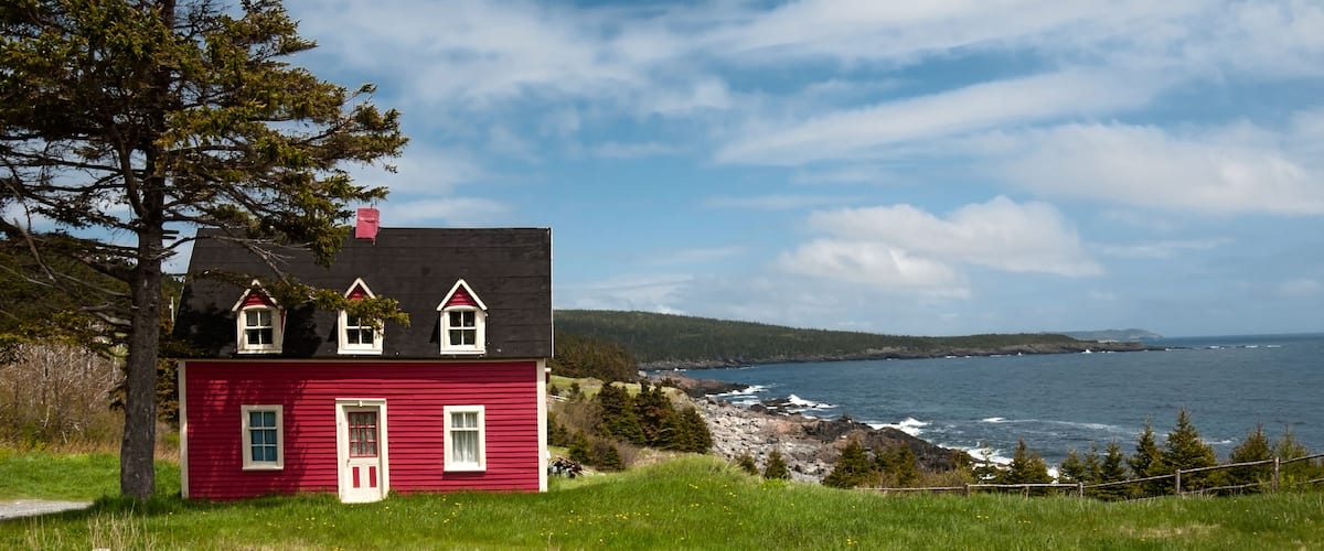 A red house with a white door and windows, a tree, and a view of the ocean and rocky coastline.