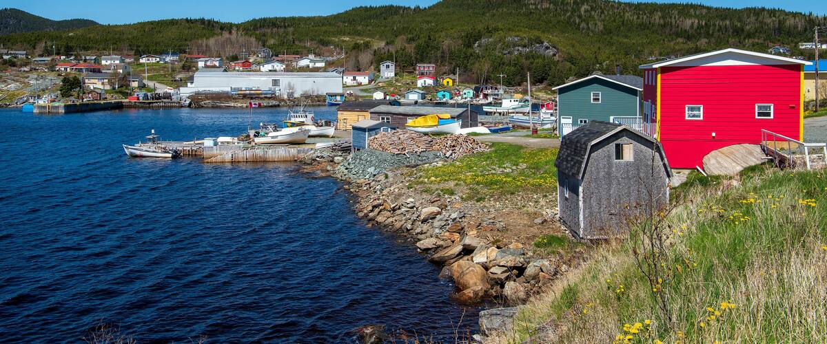 A coastal village with boats docked at a pier, a red building, and a forested hill in the background.