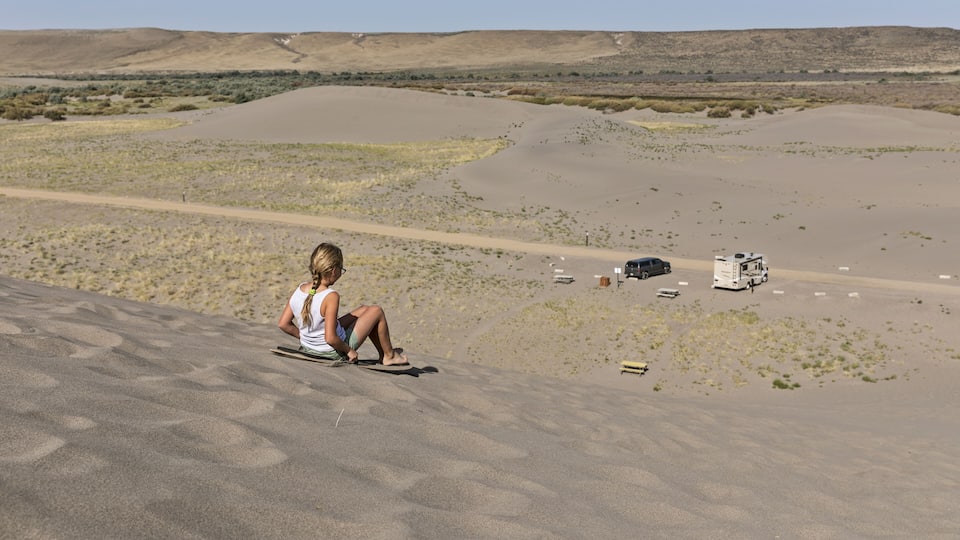 A person sitting on a sand dune overlooking a desert landscape with sparse vegetation and a road in the distance.