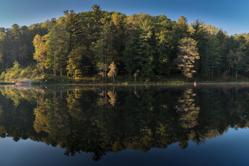 A calm lake reflecting a dense forest with a wooden bridge and a small boat.