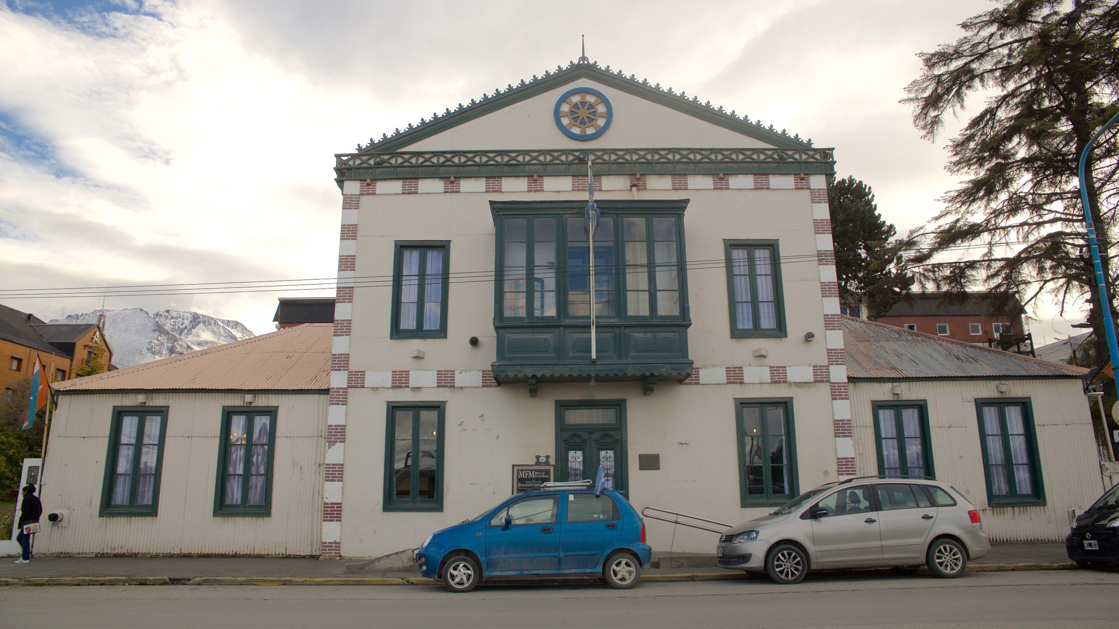 A building with a white facade and green trim, a blue car parked in front, and a mountain in the background.