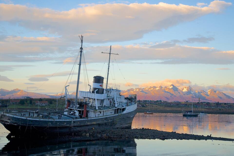 A large ship docked at a harbor with a backdrop of mountains and a sunset sky.
