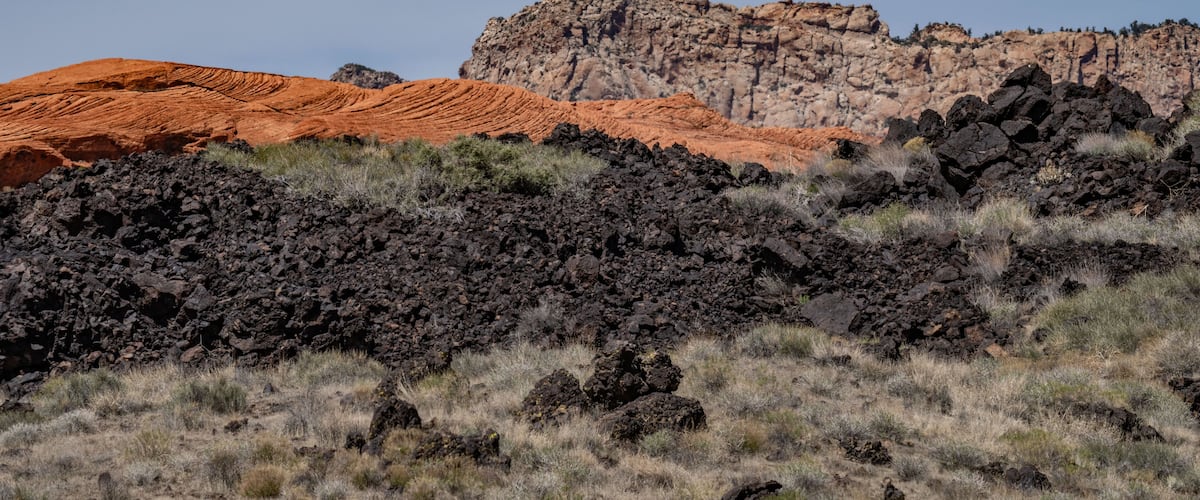 A rugged landscape with red and brown rock formations, sparse vegetation, and a clear sky.