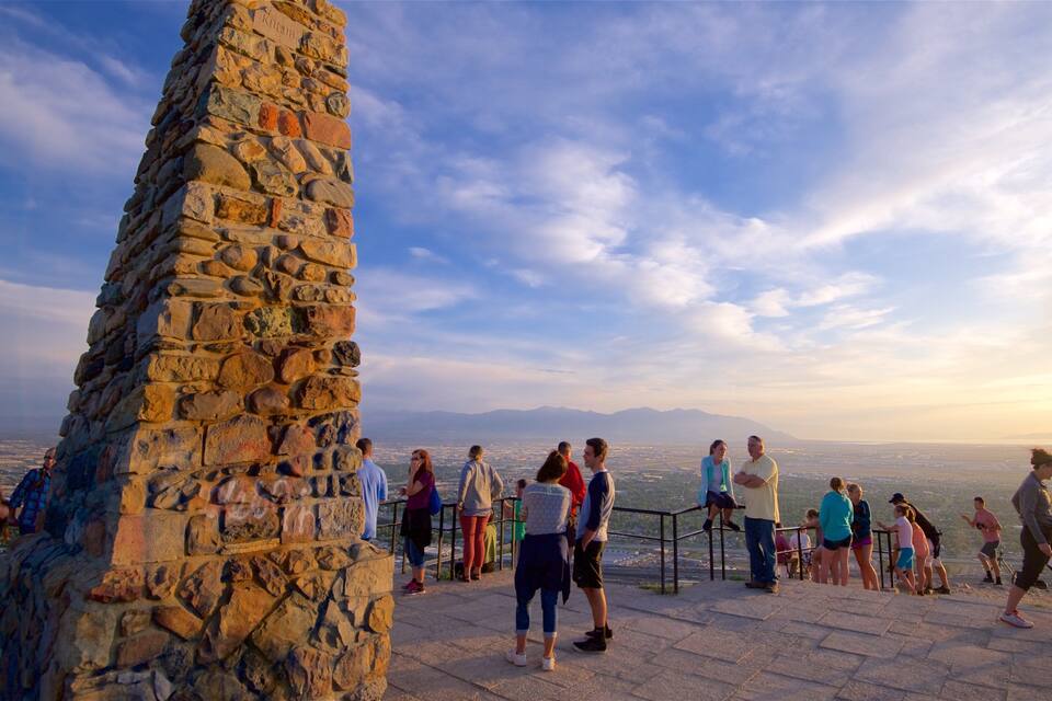 Um pilar de pedra com inscrições, um grupo de pessoas em uma plataforma de observação e um horizonte urbano ao fundo.